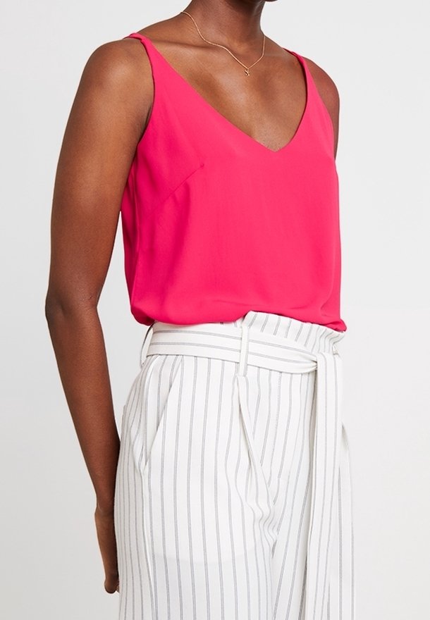 Haut rose vif sans manches avec un col en V, en tissu lisse. Associé à un pantalon blanc à fines rayures verticales et une taille nouée.