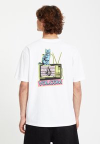 T-shirt en coton blanc avec un graphisme coloré d'un chat bleu sur une télévision vintage, portant le texte "VOLCOM" avec des accents rose et vert.