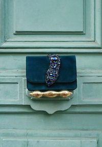 Pochette en velours bleu sarcelle avec une poignée dorée, présentant un design ondulé orné de strass bleu foncé sur le rabat.
