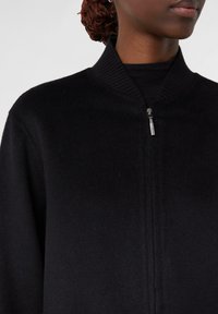 Schwarze Zip-Jacke aus weichem Stoff mit geripptem Kragen und Reißverschlussdetails, die eine glatte Textur und ein minimalistisches Design zeigt.