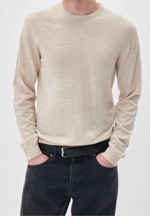 Man draagt een beige trui met lange mouwen, ingestopt in een zwarte spijkerbroek met een zwarte riem, handen in de zakken, tegen een effen achtergrond.