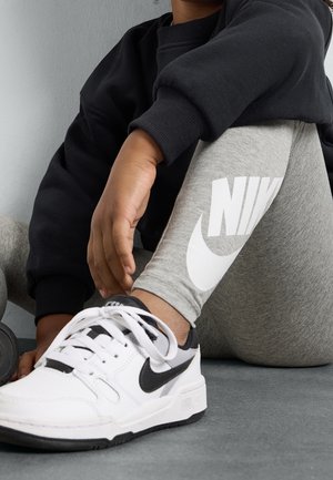 Personne portant un legging Nike gris et des baskets Nike blanches, assise avec la main posée sur le genou, vêtue d'un sweat-shirt noir.