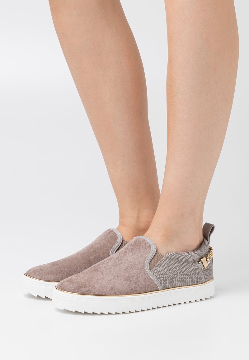 Paire de baskets slip-on taupe avec semelles blanches rainurées et détail en tissu texturé portées pieds nus sur fond blanc.