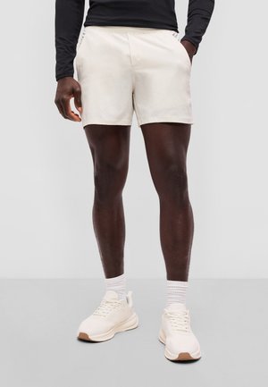 Man draagt witte sportshorts, witte sneakers met rubberen zolen, witte sokken en een zwart shirt met lange mouwen, staand tegen een effen achtergrond.