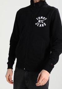 Sudadera negra con cremallera fabricada en tela suave, con el logo blanco "TOMMY NYC JEANS" en el pecho y dos bolsillos frontales.