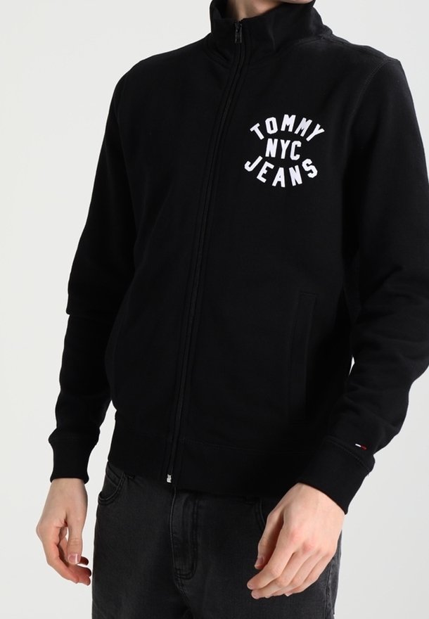 Sudadera negra con cremallera fabricada en tela suave, con el logo blanco "TOMMY NYC JEANS" en el pecho y dos bolsillos frontales.
