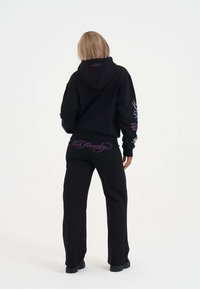 Zwart hoodie- en trainingsbroekset met een paarse Ed Hardy-logo op de achterkant. Het materiaal lijkt zacht, met een ontspannen ontwerp en lange mouwen.