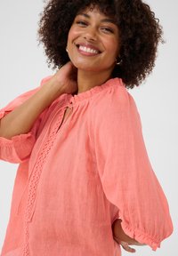 Blusa di lino corallo con scollatura rotonda, chiusura a lacci e cuciture verticali testurizzate. Presenta maniche a palloncino con polsini elasticizzati.