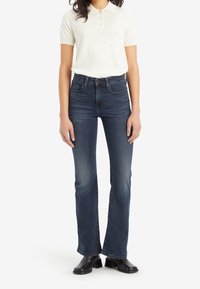 Jean bleu foncé évasé à taille haute, associé à un polo crème à manches courtes avec trois boutons. Bottines noires à la cheville complètent la tenue.