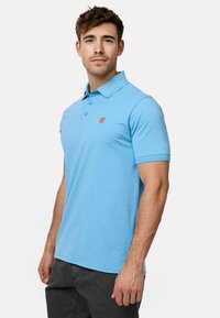 Svijetloplava polo majica od mekog materijala, s ovratnikom, tri gumba na prednjoj strani i malim narančastim logotipom na prsima.