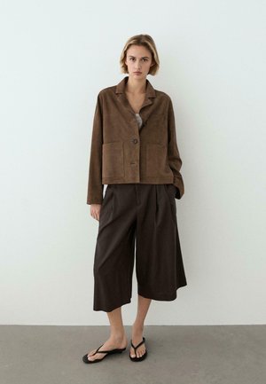 Žena stojící u bílé zdi, oblečená v hnědém semišovém saku, širokých nakrátko střižených tmavých kalhotách a černých plochých sandálech, čelem dopředu s neutrálním výrazem.