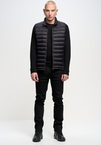 Gilet stile piumino nero con cuciture orizzontali, abbinato a una maglietta nera a maniche lunghe e jeans neri aderenti. Visibili sneakers nere e accessori.