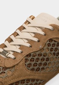 Gros plan sur une chaussure de sport en daim beige avec un motif en nid d'abeille en maille beige et des lacets blancs passés à travers des œillets en daim.