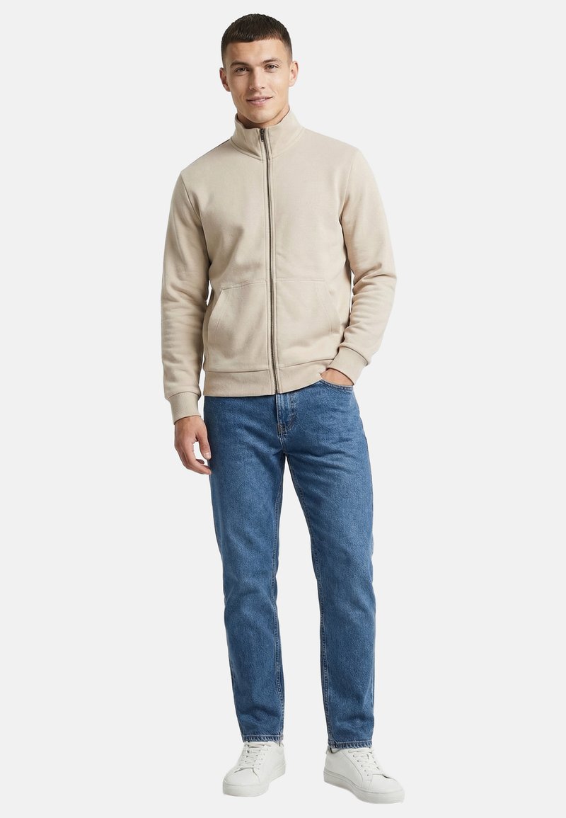 Jeune homme debout, une main dans la poche, portant une veste beige zippée, un jean bleu et des baskets blanches sur fond blanc.