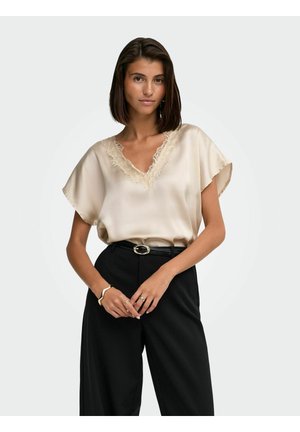 Femme portant un chemisier en satin beige avec bordure en dentelle et un pantalon noir taille haute, posant les mains jointes devant elle sur un fond clair.