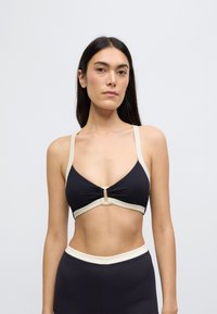 Μαύρο ριπ bralette με μπεζ ράντες και λεπτομέρεια κλεισίματος στο μπροστινό μέρος. Το ύφασμα έχει υφή που συμπληρώνει τον μινιμαλιστικό σχεδιασμό.