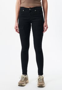 Czarne wąskie spodnie cargo z rozciągliwego materiału, z dwoma przednimi kieszeniami i eleganckim wyglądem. Połączone z beżowymi trampkami.