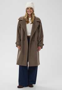 Lange bruine wollen jas met een brede kraag, dubbele rij knopen en grote zakken, gecombineerd met een crèmekleurige trui en donkerblauwe jeans met wijd uitlopende pijpen.