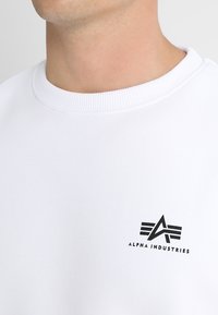 Hvid sweatshirt med ribbet rund hals og et sort trykt logo med tre linjer og teksten "ALPHA INDUSTRIES" på venstre bryst.