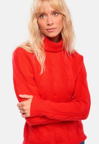 Pull en maille rouge avec un col haut et des manches longues, présentant des poignets côtelés et un motif texturé sur l'ensemble.