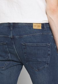Vaqueros de denim en azul oscuro con un ajuste de cintura media. Presentan bolsillos traseros y una etiqueta de marca de cuero marrón, cosida con hilo de contraste.
