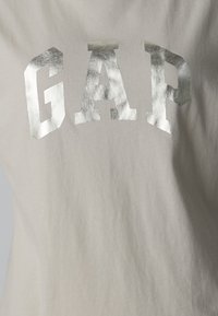 Balta medvilninė marškinėlė su didelėmis sidabrinėmis metalinėmis "GAP" raidėmis. Audinys yra minkštas, o dizainas yra paprastas, be papildomų raštų.