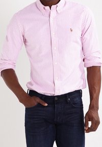 Różowa i biała w paski koszula zapinana na guziki z kołnierzykiem, z długimi rękawami podwiniętymi, w połączeniu z ciemnoniebieskimi dżinsami, z małym logo na piersi.