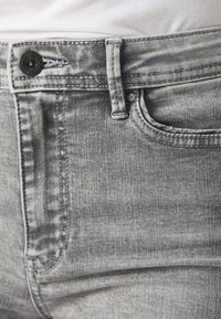 Gros plan sur la taille de jeans en denim gris avec bouton, passant de ceinture et poche avant, portés avec une chemise blanche rentrée.