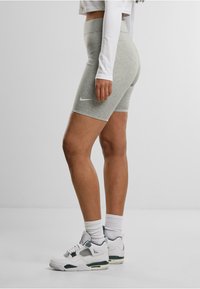 Donna che indossa pantaloni corti Nike grigi, un crop top bianco a maniche lunghe, calze bianche e sneakers bianche, in piedi contro uno sfondo neutro.