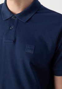 Gros plan d'une personne portant un polo bleu marine avec une patte à deux boutons et un discret écusson carré sur la poitrine.