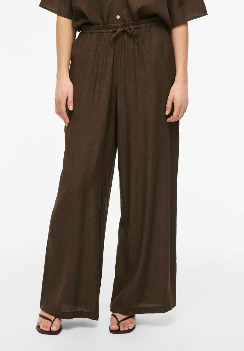 Pantalon ample marron foncé à jambes larges avec taille élastique et cordon, porté avec des sandales noires à brides sur une personne debout.