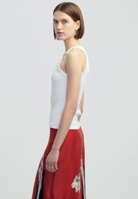 Witte geribbelde tanktop met dunne bandjes, gecombineerd met een rode rok met een bloemenpatroon. Achtergrond is lichtgrijs.