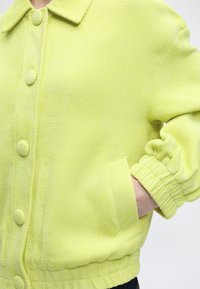 Κοντινό πλάνο ενός ατόμου που φοράει μια υφασμάτινη ανοιχτοπράσινη ζακέτα με κουμπιά καλυμμένα με ύφασμα και ένα χέρι στην μπροστινή τσέπη.