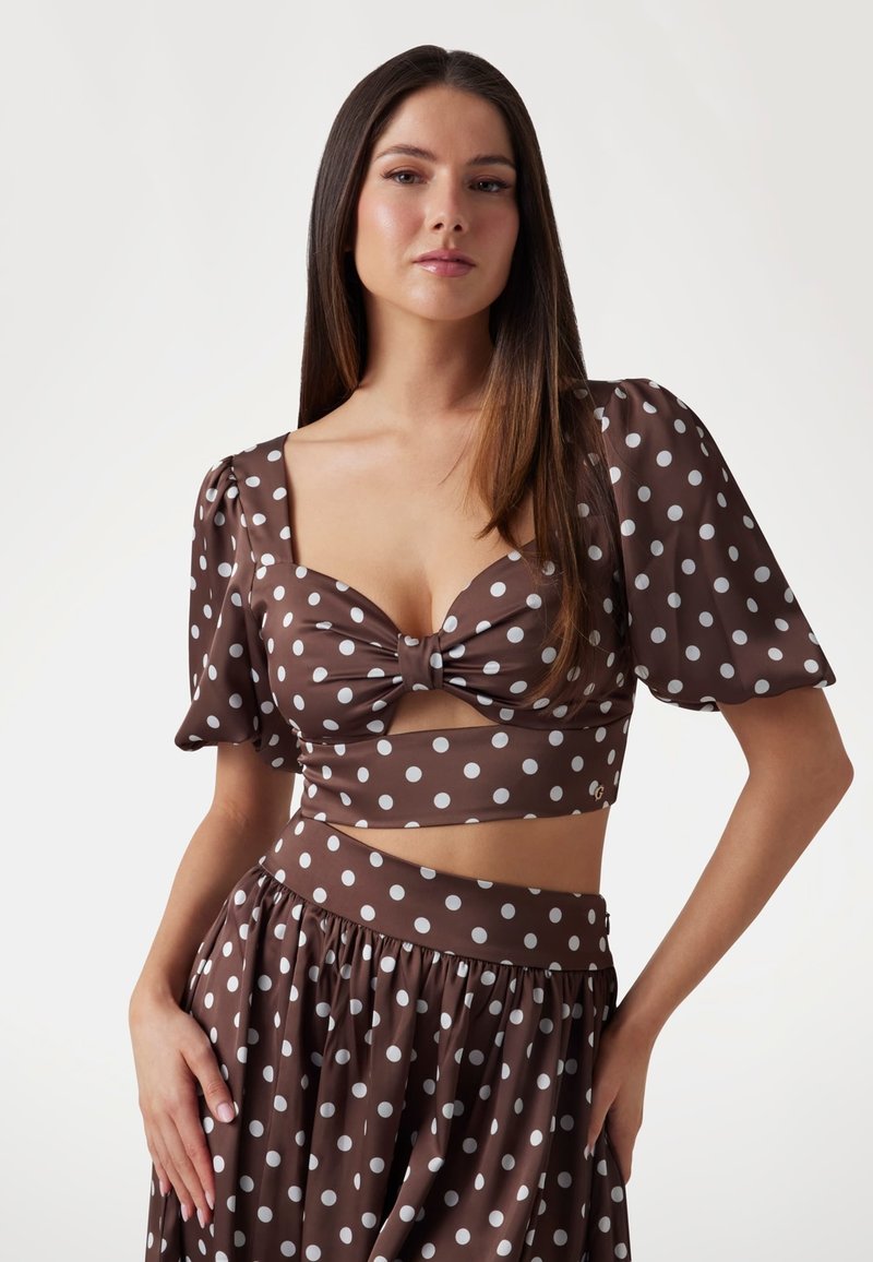 Brązowy crop top w kropki z bufkami, detal w postaci kokardy z przodu oraz pasująca spódnica o wysokim stanie. Materiał jest gładki i lekki.