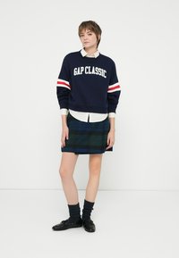 Sudadera azul marino con texto blanco "GAP CLASSIC", rayas rojas y blancas en las mangas, superpuesta sobre una camisa blanca. Falda de cuadros verde, calcetas oscuras y zapatos negros.