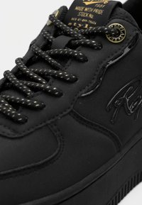 Μαύρο αθλητικό παπούτσι με χοντρές κορδόνια με σχέδιο, ανάγλυφο λογότυπο της μάρκας στο πλάι και μεταλλικό μάτι με λογότυπο κοντά στην επένδυση της γλώσσας.