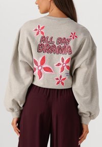 Lichtgrijze sweatshirt met ballonsleeves, voorzien van roze bloemenpatronen en de tekst "ALL DAY DRAMA" op de achterkant. Het heeft een losse fit. Donkere broek zichtbaar.