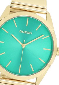 Montre en métal doré avec un cadran turquoise mat. Les marqueurs des heures et les aiguilles sont dorés. Logo "OOZOO" positionné au-dessus du centre. Design de boîtier circulaire.