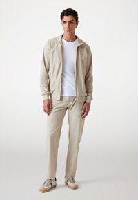 Lys beige træningsdragt med en lynlåslukning jakke og matchende bukser. Glat tekstur med diskrete syninger. Casual stil med sportssko.