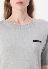 Maglione a maglia grigio chiaro con collo a giro ribattuto e maniche corte, con una piccola etichetta nera con la scritta "BLANCHE" sulla parte anteriore.
