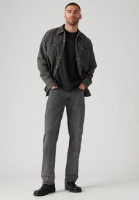 Chaqueta de mezclilla gris sobre una camiseta negra, combinada con jeans grises y botas negras. Incluye dos bolsillos en el pecho y un ajuste relajado.