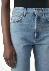 Personne portant un jean en denim bleu clair taille haute et un top noir, main détendue le long du corps sur un fond blanc uni.