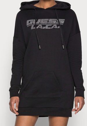 Personne portant une robe sweat à capuche noire avec une poche à l'avant et le logo "GUESS LA / CA" en points blancs sur la poitrine.