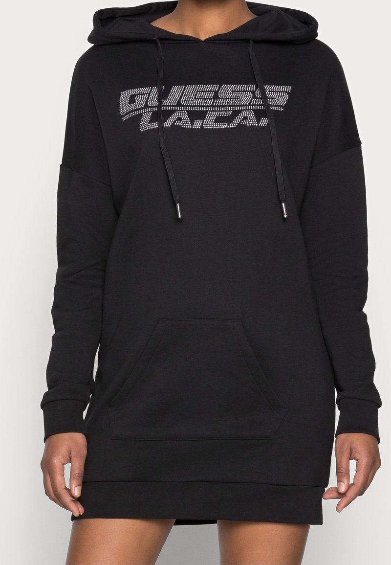 Personne portant une robe sweat à capuche noire avec une poche à l'avant et le logo "GUESS LA / CA" en points blancs sur la poitrine.