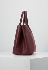 Bordeauxfarvet lædertaske med to håndtag, glat tekstur, struktureret form og diskret kornmønster. Har metalbeslag ved bunden.