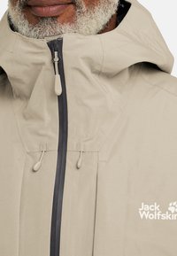 Veste imperméable beige avec un col haut, des cordons élastiques et une fermeture éclair contrastante, arborant le logo Jack Wolfskin en blanc.