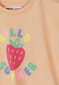 Dječja košulja boje breskve s crvenom ilustracijom jagode i šarenim slovima koja tvore riječ "Hello Summer" oko nje.