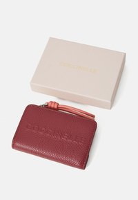 Portafoglio in pelle rossa con finitura textured, chiusura zip e un tirante della zip in pelle rosa. Presentato in una scatola rosa chiaro con branding dorato.