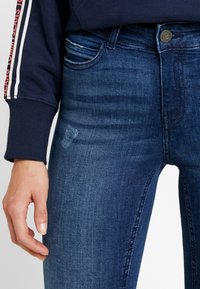 Jeans ajustados en denim de lavado oscuro con una textura suave. Presenta un bolsillo delantero, un cierre de botón metálico y detalles sutiles de desgaste.