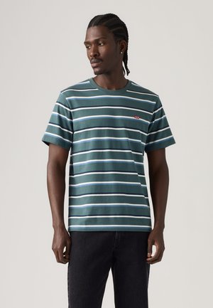 Homme aux cheveux tressés portant un t-shirt à manches courtes rayé vert et bleu et un pantalon noir, debout devant un fond uni.